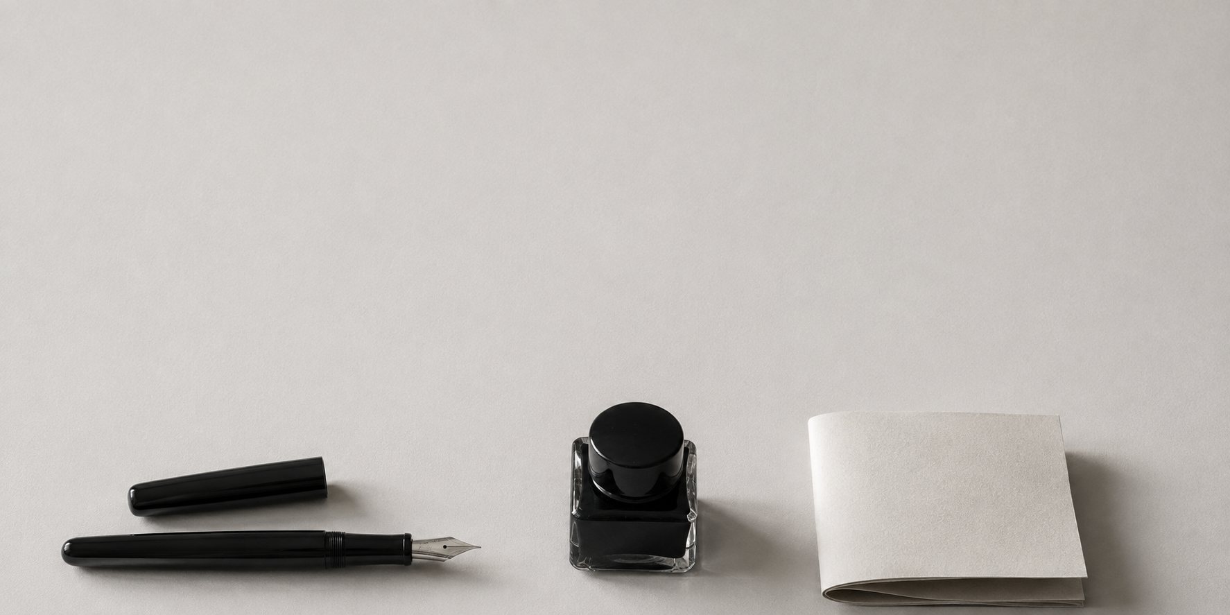 A fountain pen, a bottle of black ink and a folded sheet of paper on a plain pale desk.